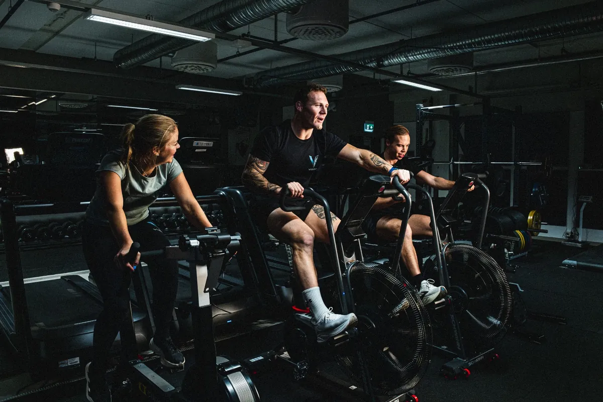 Tre personer som trener hardt på air bikes i et mørkt treningssenter