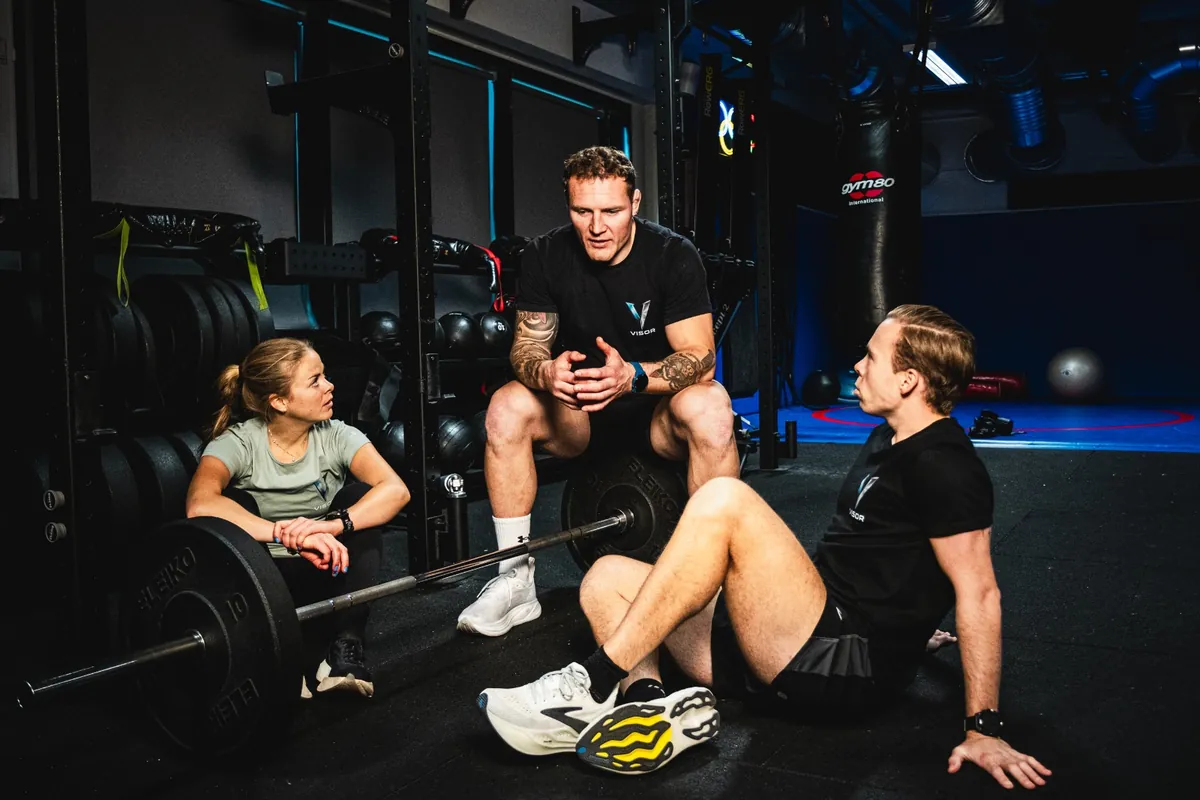 Three people sitting on a gym floor around a loaded barbell, talking through a session